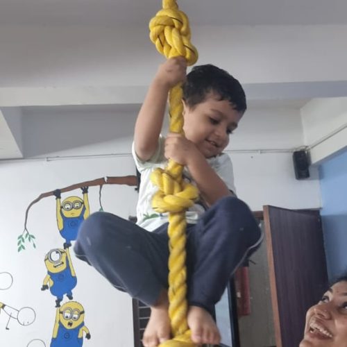 Young child climbing a yellow rope with assistance from an adult, showcasing preschool activities in Ahmedabad, with playful minion drawings on the wall.